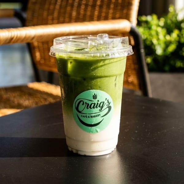 Iced matcha latte from Craig's Cafe & Bakery on a black table with a wicker chair.