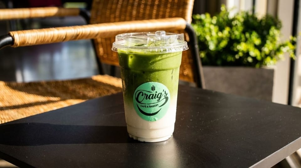 Iced matcha latte from Craig's Cafe & Bakery on a black table with a wicker chair.