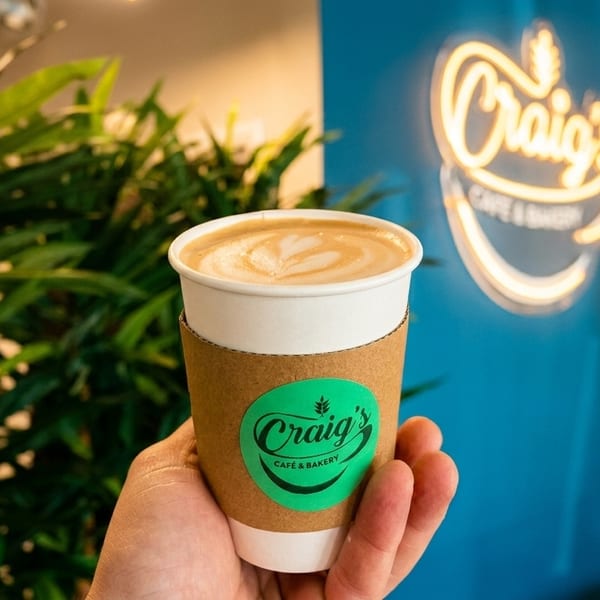 Hand holding a latte with a "Craig's Cafe & Bakery" logo, against a neon sign and green plants.